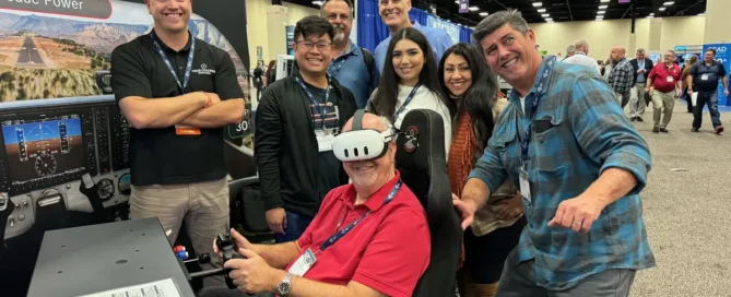 Group at a True Course Simulations expo booth, with a seated attendee wearing a VR headset flying a training device, smiling team gathered around.